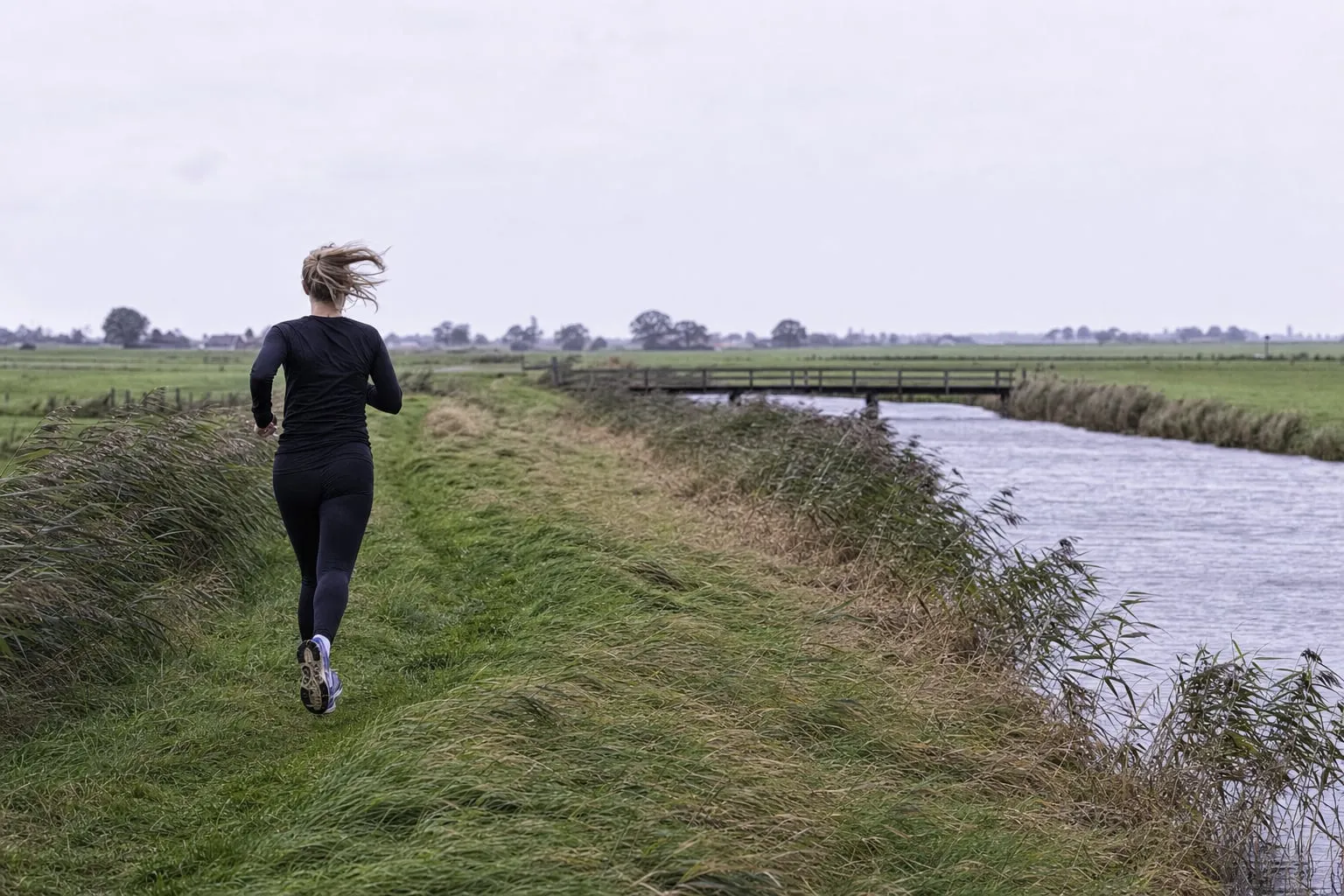 Grasbewachsener Deichweg an einem flachen Fluss, im Hintergrund eine kleine Brücke