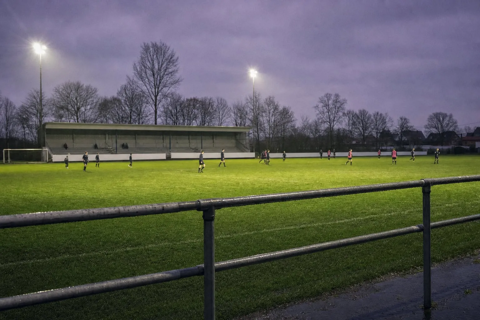 Flutlichtanlage über einem leeren Amateurfußballplatz am Abend, dahinter eine fast leere Tribüne